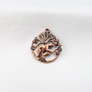 May include: A circular copper-toned metal pendant featuring an openwork design of a bear walking along a tree branch. The charm has a small loop at the top for attachment and displays a detailed, textured relief of the animal and foliage.