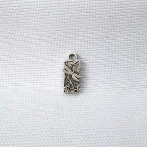 Peut inclure: Une breloque rectangulaire en métal argenté présentant un motif de libellule en relief posée sur de fines tiges linéaires rappelant de l'herbe. La breloque possède une petite boucle circulaire au sommet et présente une finition oxydée vieillie.