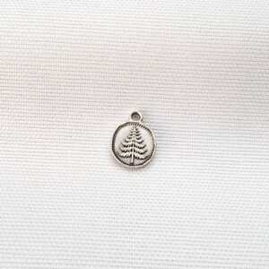 May include: A small, circular silver-toned metal charm featuring an embossed evergreen tree design in the center. The charm has a smooth, raised border and a small loop at the top for attachment, presenting a metallic finish with dark detailing.