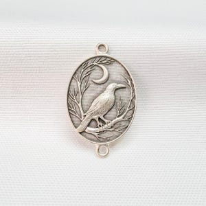 May include: An oval silver-toned metal pendant featuring a raised relief design of a bird perched on a branch with a crescent moon above. The piece has a small circular loop at both the top and bottom for attachment, with a textured background.