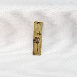 May include: A rectangular brass-toned metal pendant featuring a stamped dandelion seed head design. The charm has a textured, weathered finish and includes a single circular hole at the top for attachment, presenting a flat, elongated shape.