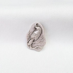 May include: A silver-toned metal pendant with an irregular, organic shape featuring a raised relief design of a bird perched on a branch. The surface has a matte, antiqued finish with dark oxidation in the recessed areas to highlight the details.