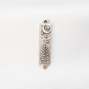 May include: A rectangular silver-toned metal jewelry connector charm featuring an embossed design of a crescent moon, multiple small stars, and a single fern leaf. The charm has a small circular loop at both the top and bottom ends.