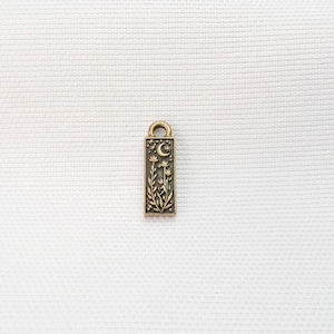 May include: A rectangular bronze-toned metal charm featuring an embossed design of a crescent moon, small stars, and a cluster of wildflowers. The charm has a small circular loop at the top for attachment and a dark, recessed background finish.