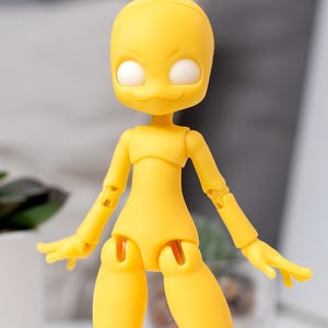 May include: A yellow articulated doll figure with a smooth matte finish, featuring a rounded head, large white circular eyes, and a small mouth. The figure has jointed shoulders, elbows, hips, and knees, allowing for various poses.