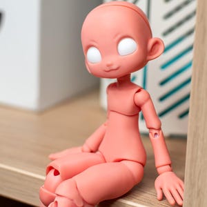 May include: A peach-colored ball-jointed doll with a smooth, matte finish. The doll features a rounded head, large blank white eyes, and a small, neutral mouth. Its articulated body is posed in a seated position with visible joints at the shoulders, elbows, hips, and knees.