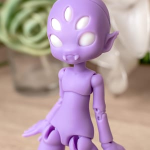 以下が含まれることがあります： ラベンダー色の球体関節人形。4つの白い目と2本の小さな牙を持つ独特な頭部が特徴です。尖った耳、滑らかなマット仕上げの表面を持ち、首、肩、腰に関節があるため、様々なポーズをとることが可能です。