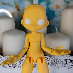 May include: A yellow articulated ball-jointed doll with a smooth, matte finish. The doll features a rounded head, large blank white circular eyes, and a simple facial structure. Its body has visible joints at the shoulders, elbows, and hips.