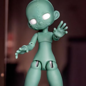 May include: A pale mint green articulated doll with a smooth matte finish. The doll features a vertical stitched seam detail running down the center of its face and glowing white circular eyes. Its limbs are jointed, and it is posed with one hand raised.