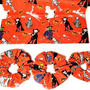 May include: Three fabric hair scrunchies and a rectangular piece of fabric featuring an orange background with a repeating pattern of Halloween characters including skeletons, ghosts, vampires, witches, and jack-o'-lanterns in various colors.