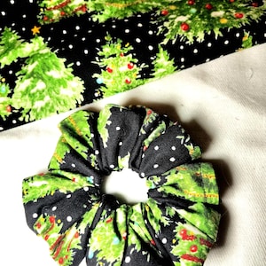May include: A fabric hair scrunchie featuring a black background with a pattern of green Christmas trees decorated with red and yellow ornaments, accented by small white dots resembling falling snow throughout the textile design.