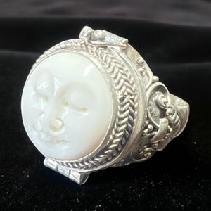 May include: A silver ring featuring a carved white moon face. The ring has an ornate, detailed design with a braided border around the moon and decorative elements on the sides. The ring is set against a dark background.