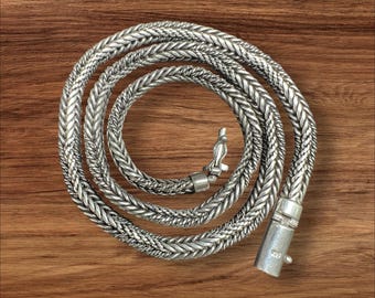 Puede incluir: Un collar de cadena de plata con un diseño trenzado. La cadena está enrollada sobre una superficie de madera.