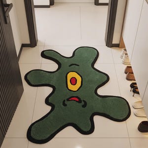 May include: An irregular, amoeba-shaped rug in a deep olive green color with a thick black border. The rug features a central yellow oval eye with a red pupil and a small, curved red mouth below it, creating a cartoon-like character design.