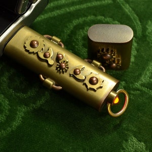 May include: A steampunk-themed USB flash drive and case. The drive and case are brass-toned, decorated with gear-shaped details and copper elements. The drive features a small, illuminated orange light at the end. The items are on a green surface.