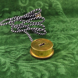 Pode incluir: Um pingente circular de cor latão com um design de buraco de fechadura e uma borda texturizada. Está preso a um cordão trançado em preto e branco. O pingente está sobre um tecido verde estampado.