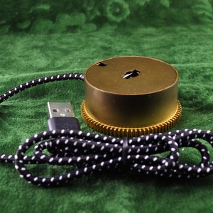 Pode incluir: Um objeto cilíndrico de cor latão com uma base em forma de engrenagem e uma pequena abertura na parte superior. Um cabo USB preto com bolinhas brancas está conectado ao objeto. O fundo é um tecido verde texturizado.