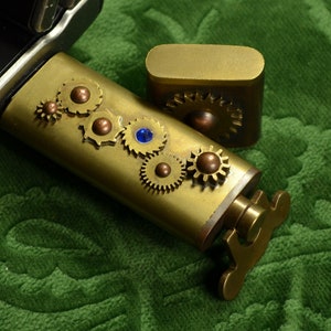 Può includere: Un accendino in ottone in stile steampunk e una scatola abbinata, adornati da ingranaggi decorativi e una gemma blu. L'accendino presenta un coperchio incernierato e un meccanismo a forma di chiave. Su sfondo verde.