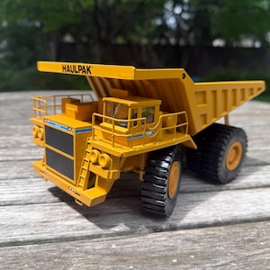 May include: A yellow Haulpak mining dump truck scale model featuring a large rear bed, a cab with railings, a front grille, and black tires. The word HAULPAK is printed in black letters on the top front edge of the truck's large yellow dump bed.