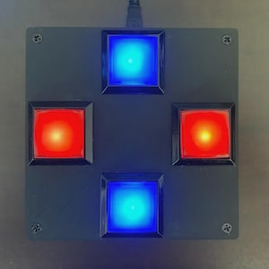 May include: A square black electronic device featuring four large, illuminated square buttons arranged in a cross pattern. The top and bottom buttons glow with a bright blue light, while the left and right buttons glow with a vibrant red light.