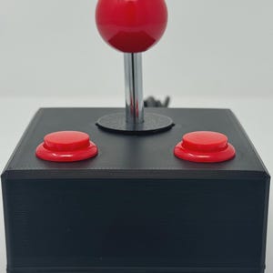 Peut inclure: Une base de contrôleur d'arcade rectangulaire noire dotée d'un joystick central avec une tête sphérique rouge brillante et une tige métallique. Deux boutons rouges circulaires sont placés symétriquement sur le dessus du boîtier noir.