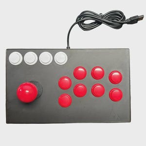 May include: A rectangular black arcade controller featuring a red joystick on the left, eight red action buttons arranged in two rows, and four white buttons along the top edge. A black USB cable is attached to the top of the device.