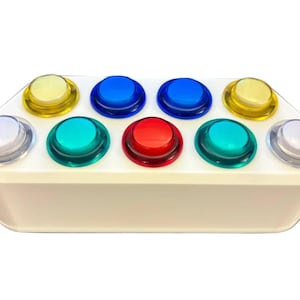 Puede incluir: Una base de controlador rectangular blanca con diez botones circulares translúcidos dispuestos en dos filas. La fila superior incluye botones amarillos y azules, mientras que la fila inferior presenta botones blancos, turquesas y rojos.
