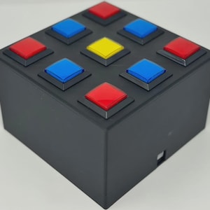 May include: A black square box featuring a three-by-three grid of nine raised buttons on its top surface. The buttons are arranged in a pattern with three red buttons, four blue buttons, and one yellow button positioned in the center.