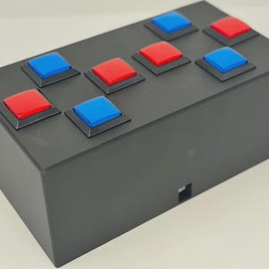 May include: A rectangular black box featuring eight square buttons arranged on the top surface. The buttons are colored in alternating red and blue, each set within a slightly raised black square frame. A small square port is visible on the side.