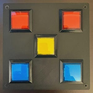 May include: A square black panel featuring five recessed square buttons arranged in a cross pattern. The top two buttons are red, the center button is yellow, and the bottom two buttons are blue. Each button is framed in black plastic.