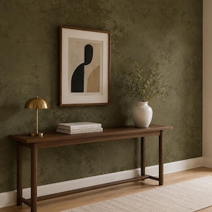 May include: A long, dark wood console table stands against a textured olive green wall. On the table sits a brass dome-shaped lamp, two stacked books, and a white ceramic vase containing green branches. A framed abstract art print hangs above.