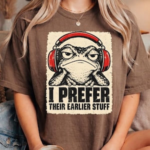 May include: A brown short-sleeve t-shirt featuring a graphic of a toad wearing red headphones. The toad rests its chin on its hands. Below the graphic, the text reads I PREFER THEIR EARLIER STUFF in a bold, distressed black font on a cream background.