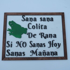 May include: A rectangular wooden sign with a dark brown frame and a white background. It features a green frog cutout on the left and black raised lettering that reads: Sana sana Colita De Rana Si NO Sanas Hoy Sanas Mañana.