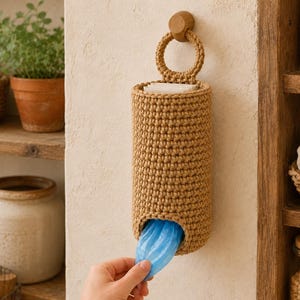 Puede incluir: Una bolsa de almacenamiento cilíndrica tejida a ganchillo en color beige, con un asa circular superior colgada de un gancho de pared de madera. La bolsa tiene una abertura inferior de la que se extrae una bolsa de plástico azul.