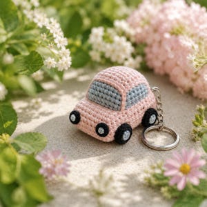 Pode incluir: Um pequeno porta-chaves de carro em croché com carroçaria rosa claro, janelas cinzentas claras e três rodas pretas visíveis com centros cinzentos claros. Uma corrente de metal prateado e um anel circular estão presos ao veículo.