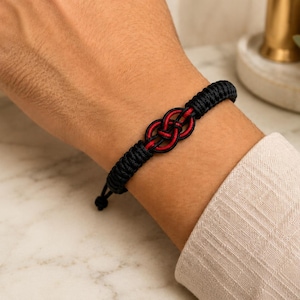 May include: A black cord bracelet featuring a central decorative knot design with interwoven red and black threads. The bracelet is constructed from braided cord and includes a sliding knot closure mechanism for adjustment on the wrist.