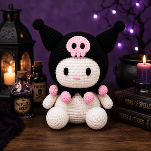 May include: A handmade crochet plush toy featuring a white body and a black hood with two pointed ears. The hood is decorated with a pink skull emblem. The character has a black collar adorned with small pink pom-poms and simple black eyes.