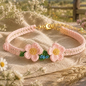 May include: A light pink crochet bracelet featuring two five-petalled flowers with yellow centres, small green leaves, and tiny blue accents. The band is made of woven pink thread and includes a gold-toned metal lobster clasp and an adjustable chain.