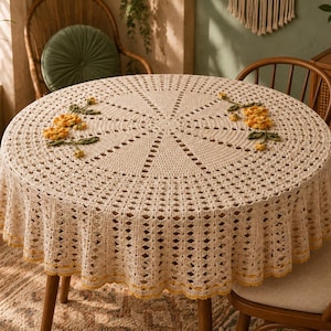 May include: A round, cream-coloured crocheted tablecloth featuring an intricate openwork pattern and a scalloped edge with a thin yellow border. Two clusters of three-dimensional yellow flowers with green leaves are attached to the top surface.