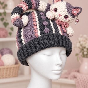 Pode incluir: Um gorro de crochê com borda canelada cinza-escura e coroa com listras verticais em tons de creme, rosa e cinza-escuro. Um gato de crochê tridimensional com grandes olhos roxos e um pequeno pingente de coração fica no topo do gorro.