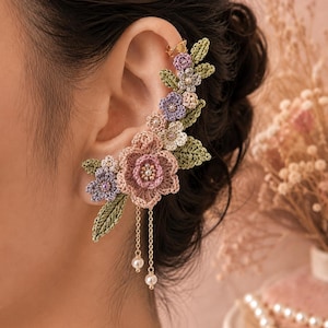Könnte beinhalten: Eine gehäkelte Ohrmanschette mit einem Blumenarrangement aus rosa, lavendelfarbenen und weißen Blüten sowie grünen Blättern. Das Schmuckstück besitzt oben eine goldfarbene Metallmanschette und zwei feine Goldketten mit jeweils einer kleinen Perle.