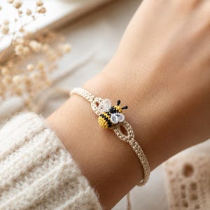 Puede incluir: Una pulsera de macramé beige que presenta un pequeño dije de abeja de ganchillo en el centro. La abeja tiene un cuerpo a rayas amarillas y negras con delicadas alas blancas similares a hilos y dos pequeñas antenas negras. La banda es de cordón fino.