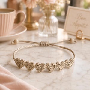 May include: A beige crochet bracelet featuring six small, heart-shaped motifs arranged in a row. The bracelet has a sliding knot closure with two small wooden beads at the ends of the adjustable cord, crafted from a light tan fiber material.