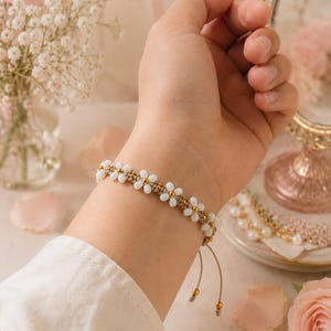 May include: A bracelet featuring a repeating daisy pattern made of small white petal-shaped beads and central gold-toned beads, strung on a light tan cord. The adjustable design includes two dangling cord ends finished with small gold-toned beads.
