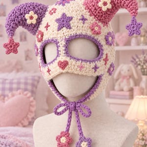 Pode incluir: Um gorro de bobo da corte em crochê na cor creme com dois chifres longos e pontiagudos em roxo e rosa. O gorro é decorado com pequenas estrelas e flores de crochê roxas e rosas, um detalhe em forma de coração e laços roxos com flores.