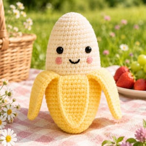 Puede incluir: Un peluche de banana tejido a ganchillo con la parte superior color crema y la cáscara de color amarillo brillante. El juguete tiene dos ojos de cuentas negras, una pequeña sonrisa negra bordada y dos mejillas circulares rosas.