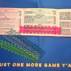 May include: A Mah Jongg score card with a pink, a clear glittery, and a green scalloped acrylic marker. The card lists various hand combinations and point values. The text at the bottom reads, "JUST ONE MORE GAME Y'ALL" in yellow letters.