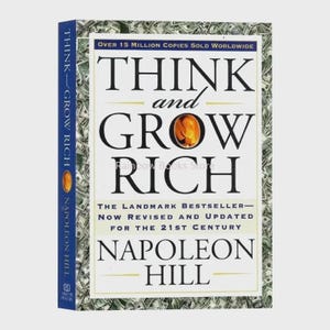 Peut inclure: Un livre relié intitulé Think and Grow Rich de Napoleon Hill. La couverture présente un fond blanc avec du texte noir, un petit graphique circulaire orange et une bordure composée de billets de banque américains. Le texte indique une édition révisée.