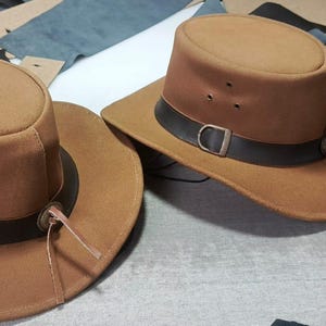May include: Two tan felt hats with flat crowns and wide brims. Each hat features a dark brown leather band around the base of the crown, secured with a metal buckle and a thin leather cord tie. Three small metal eyelets are visible on the side.