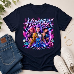 May include: A navy blue t-shirt featuring a vibrant graphic print of three anime-style characters and a blue cat. The text HUNTRIX is displayed in a bold, stylized font above the characters, surrounded by neon pink and blue lightning bolts.